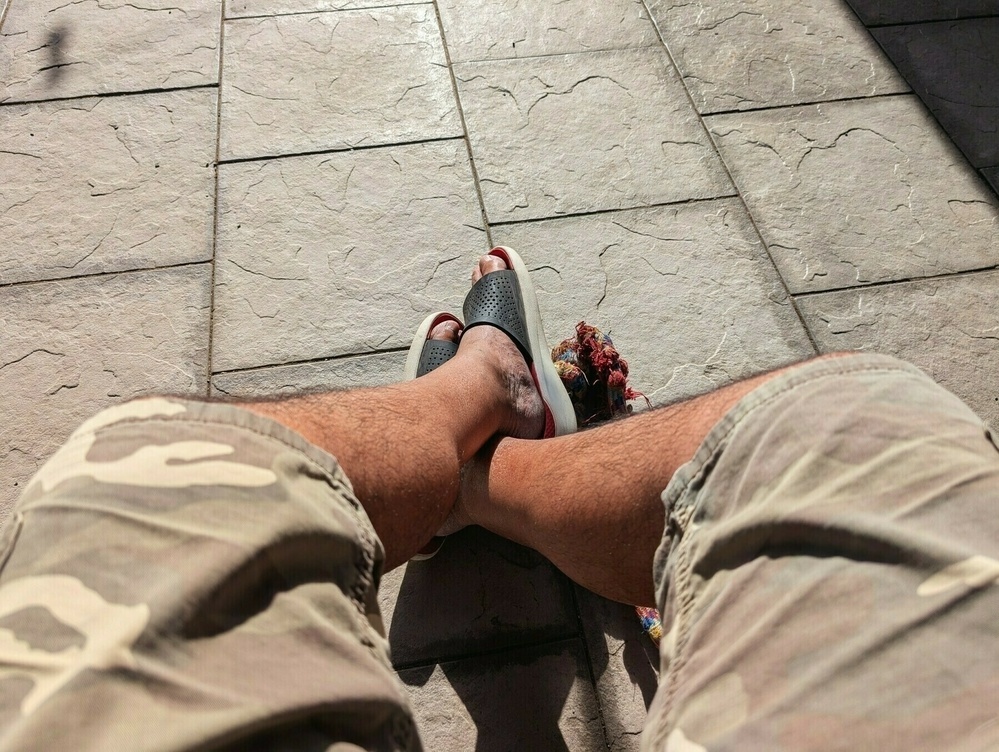 A person wearing camouflage shorts and sandals is sitting on a tiled outdoor surface, with their legs crossed.