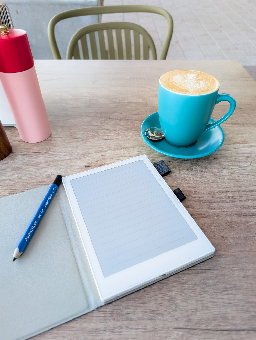 A tablet or e-reader and a pen rest on a wooden table next to a teal cup with a latte art design on top.