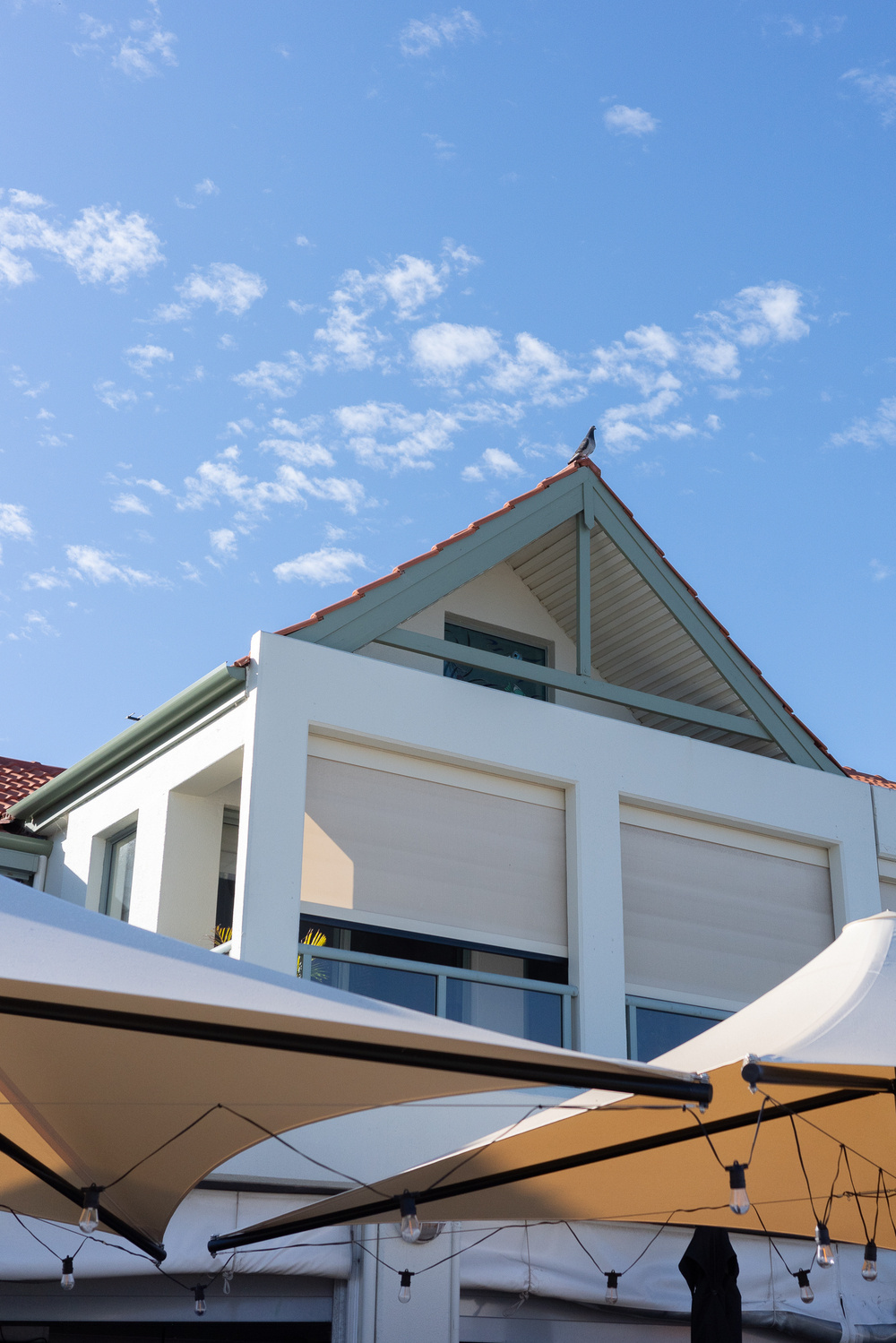 A modern house with a red-tiled roof sits under a clear blue sky, framed by patio umbrellas in the foreground.