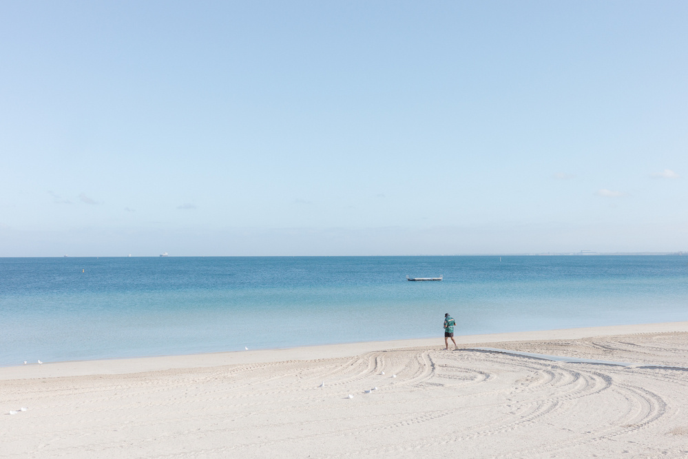 A solitary person walks along a pristine, empty beach with a small boat in the distance on the calm sea.