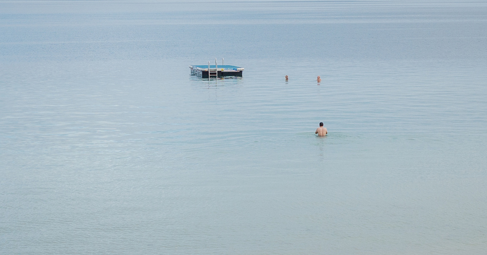 A person wades in calm, shallow water, with two more people and a floating platform further out in the distance.