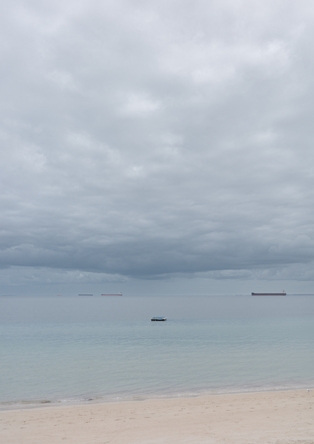 A small boat floats on a calm sea under a cloudy sky, with large ships visible in the distance.