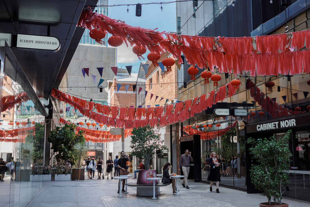 A pedestrian walkway is adorned with red lanterns and decorations, with people walking beneath and storefronts lining the path.