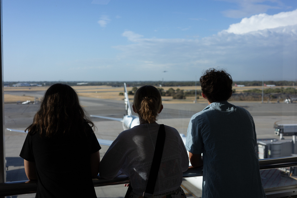 Three people are standing together, looking out at an airport runway with airplanes in the background.