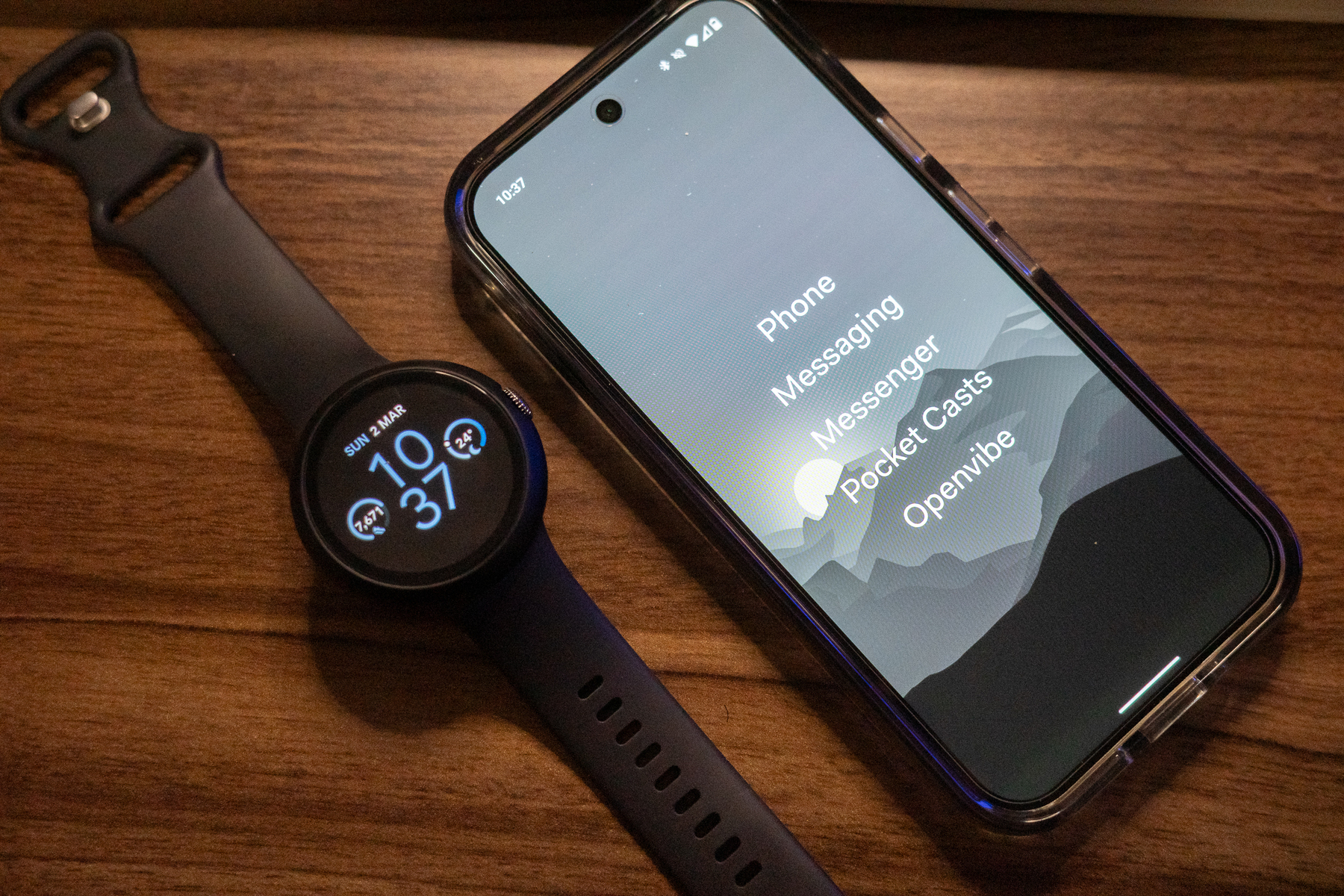 Auto-generated description: A smartwatch displaying the time sits next to a smartphone showing a list of apps on a wooden surface.