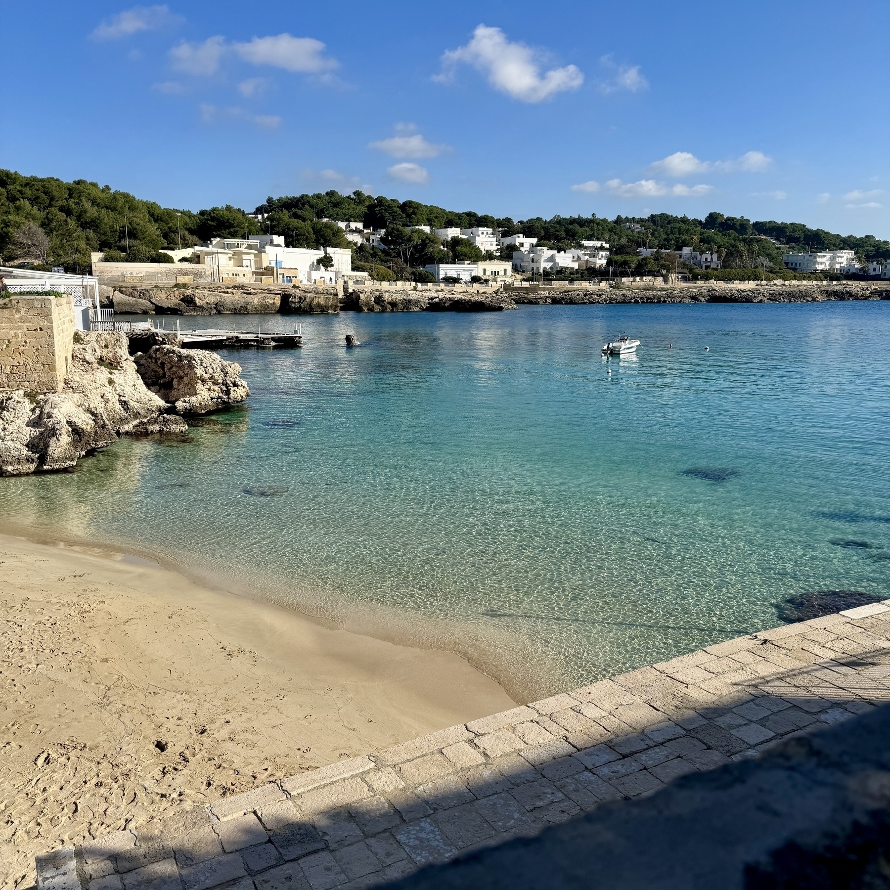 Santa Caterina beach in Nardò. You can see a sandy beach, clear blue sky and some trees on the hills. 