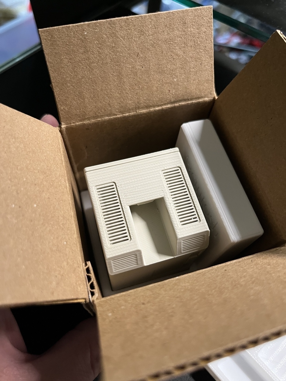A cardboard box contains a small, rectangular plastic holder with vents, revealing an open slot in the center.