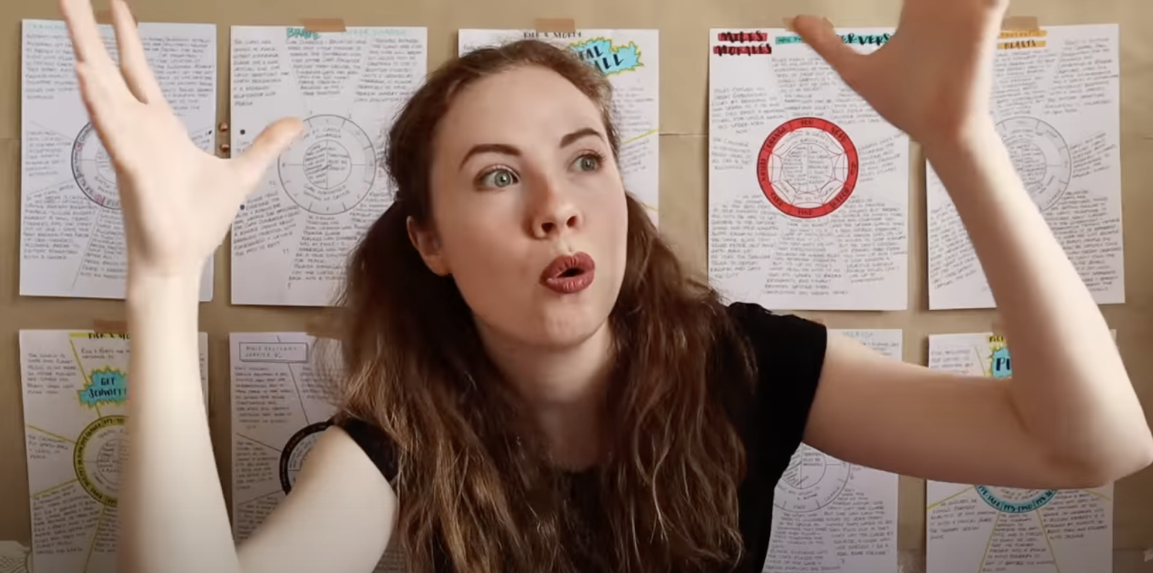 Thumbnail of Rachael Stephen video with her in front of a corkboard with several "plot embryo" papers pinned to it. Rachael with arms lifted after making a "pshew" sound for "mind blown" from video.