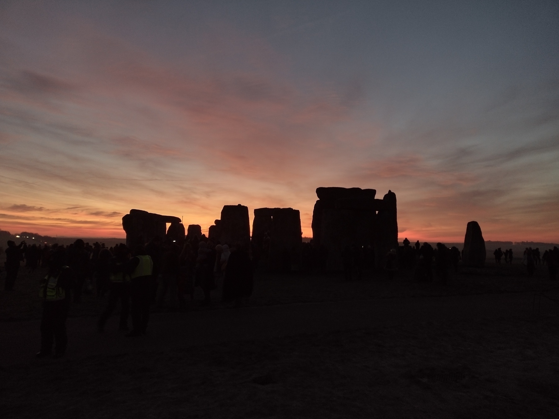 A group of people are gathered around the silhouetted stone structures of Stonehenge at sunset.