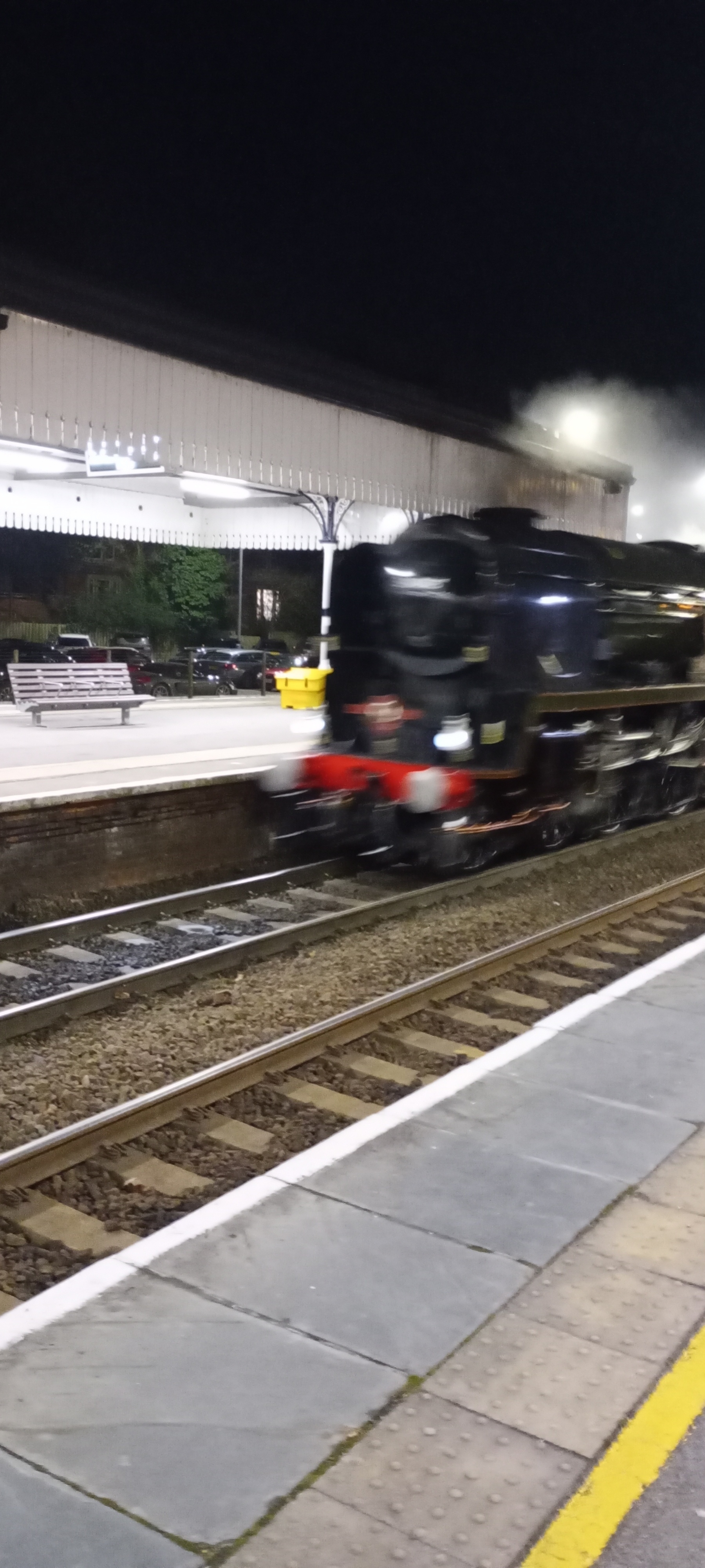 A steam train is passing through a dimly lit train station at night.