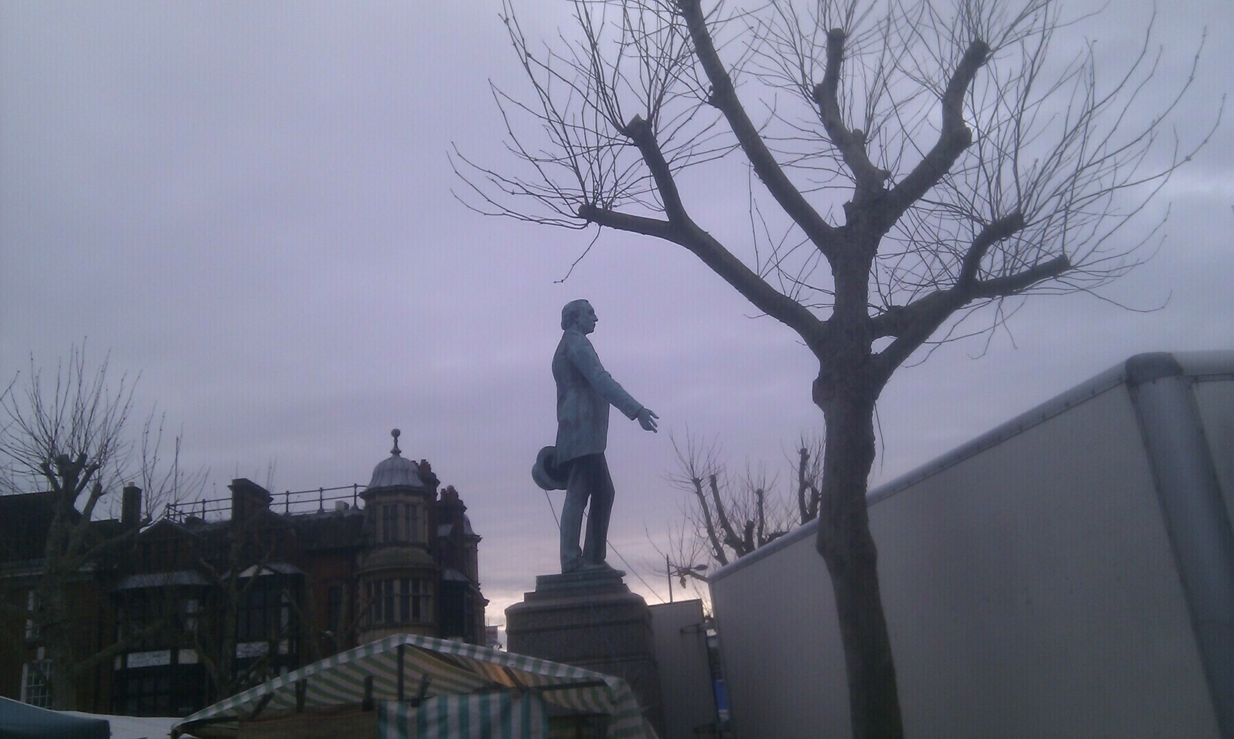 A statue of a standing figure is placed on a pedestal beside leafless trees against a cloudy sky.