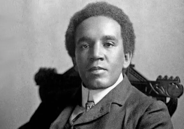 A man in a suit is seated on an ornate chair, looking directly at the camera.