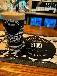 A pint of Outland Badger Stout sits on a bar mat displaying the same branding, with bottles and glasses in the blurred background.