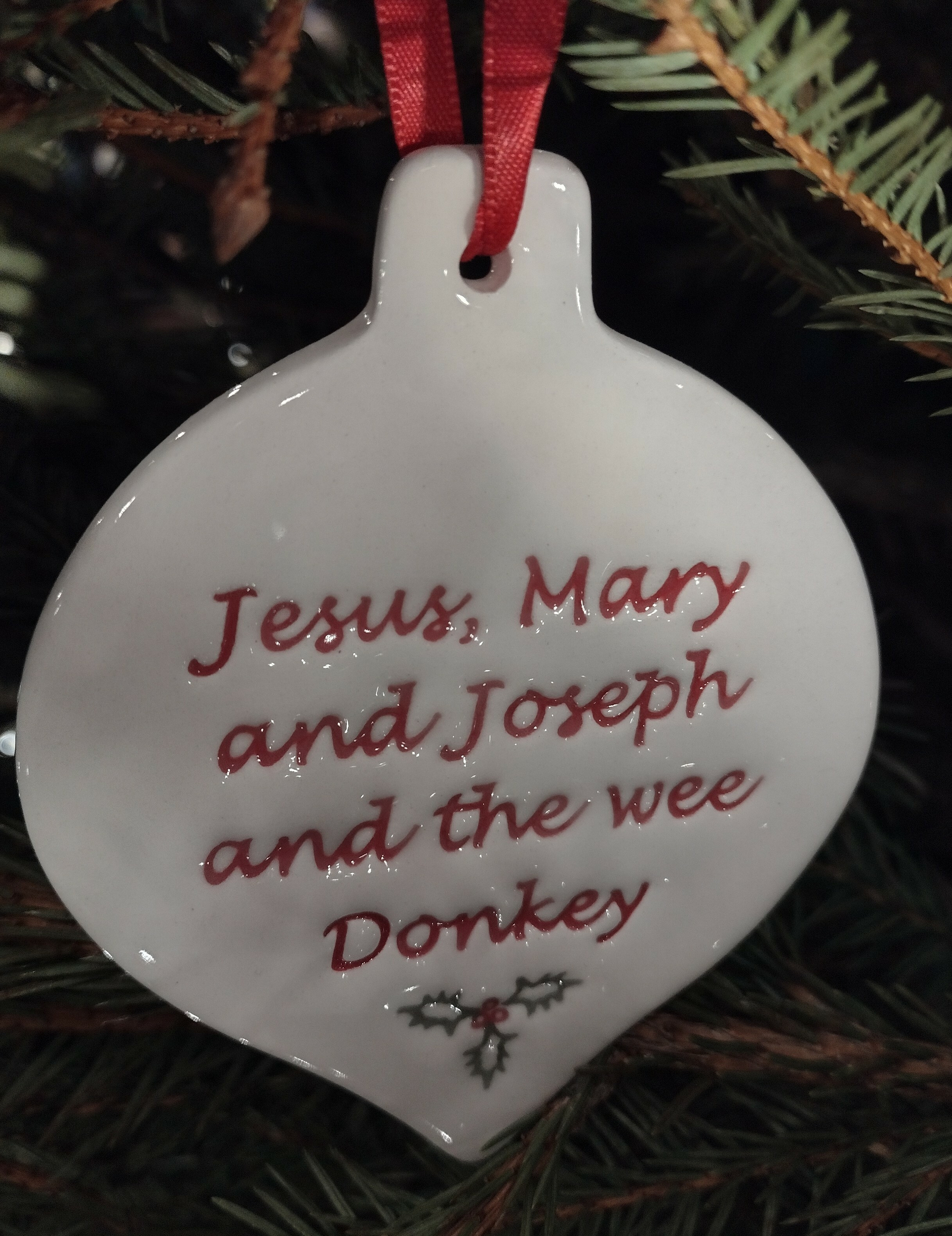 A white ornament with red text hanging on a Christmas tree reads, Jesus, Mary and Joseph and the wee Donkey, and is adorned with a sprig of holly.