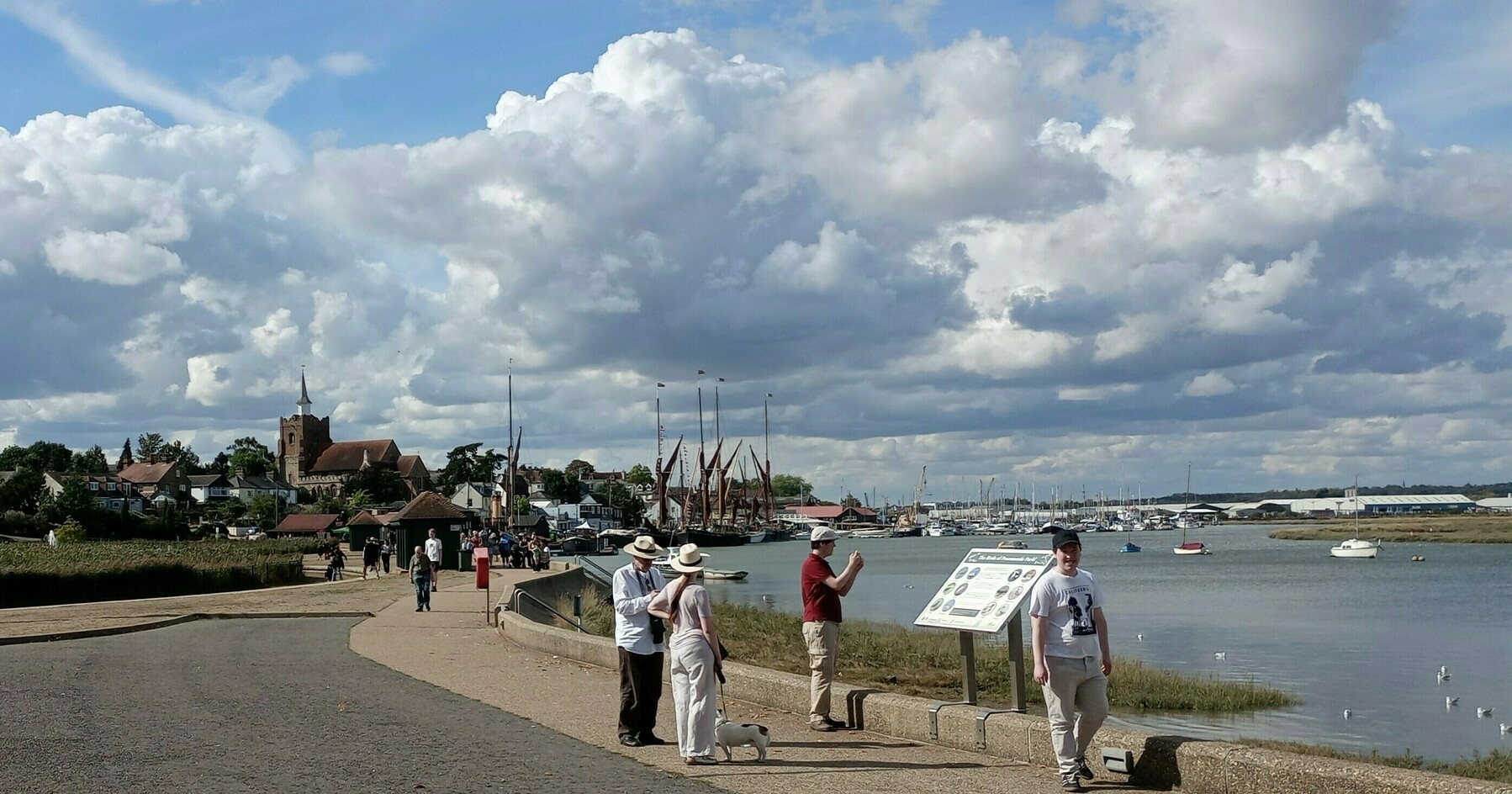 People are walking and enjoying the view along a waterfront path with boats and a village in the background under a sky filled with clouds.