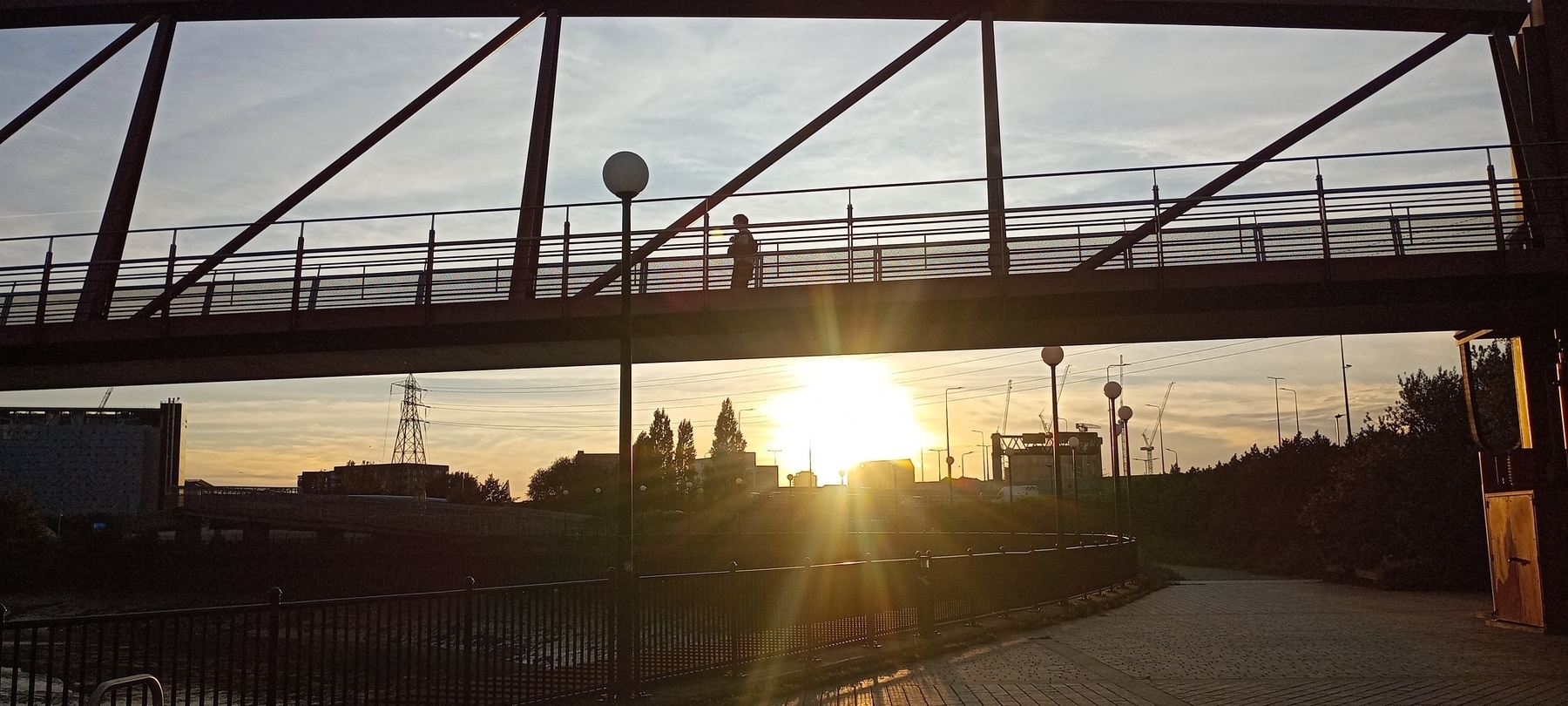 Auto-generated description: A person walks across a bridge during sunset with a scenic cityscape and river below.