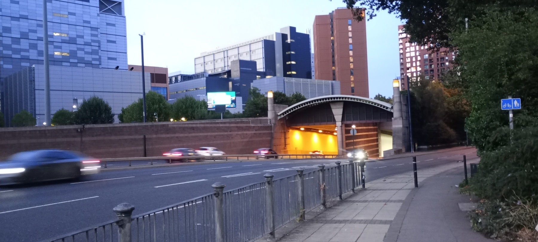 Auto-generated description: A road with light traffic passes by a modern urban setting featuring tall buildings and an illuminated tunnel entrance.