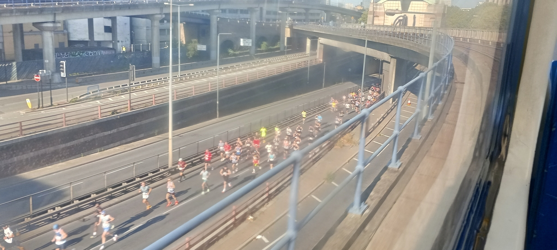 A group of people is running on a highway beneath an overpass as seen from the Docklands Light Railway 
