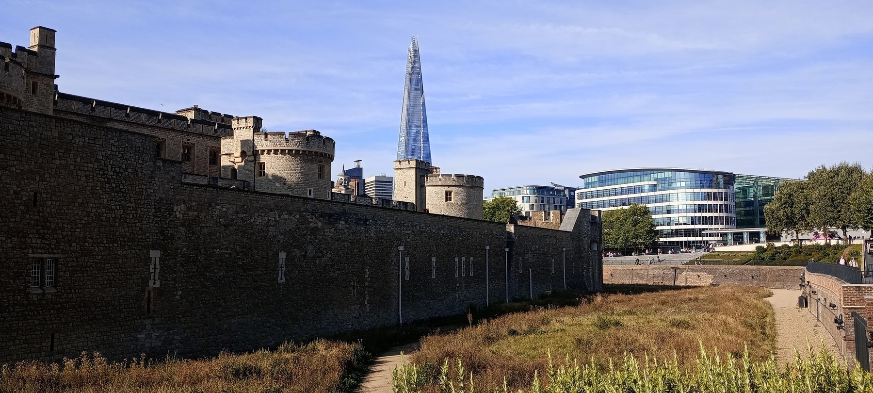 Auto-generated description: A historic stone wall with towers is in the foreground, with modern buildings, including a tall, glass skyscraper, visible in the background.