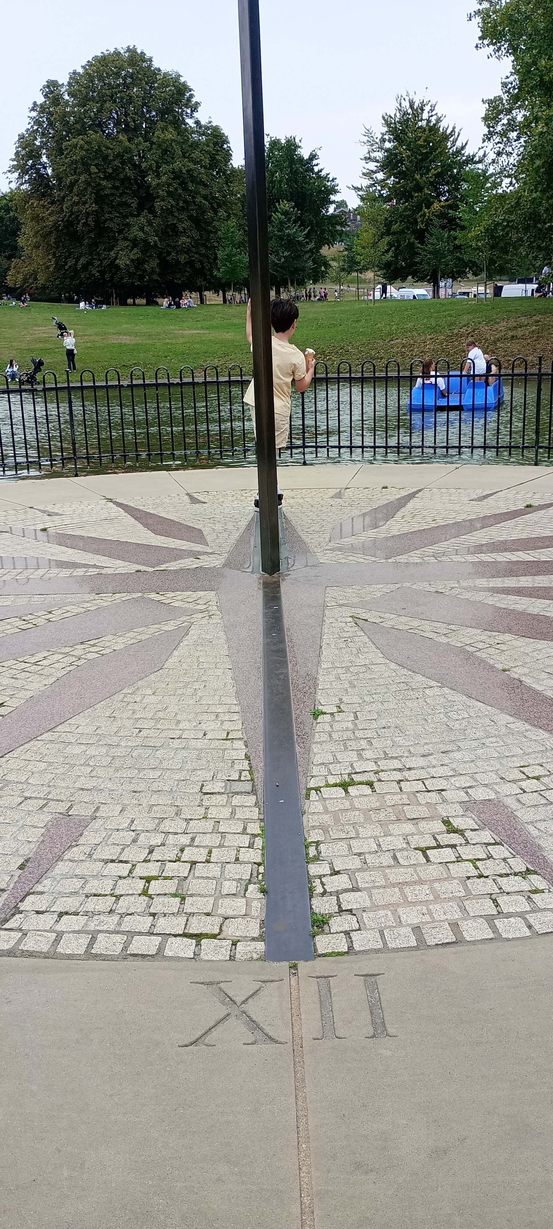 Auto-generated description: A sundial from XII is surrounded by trees, water, and people enjoying a park.