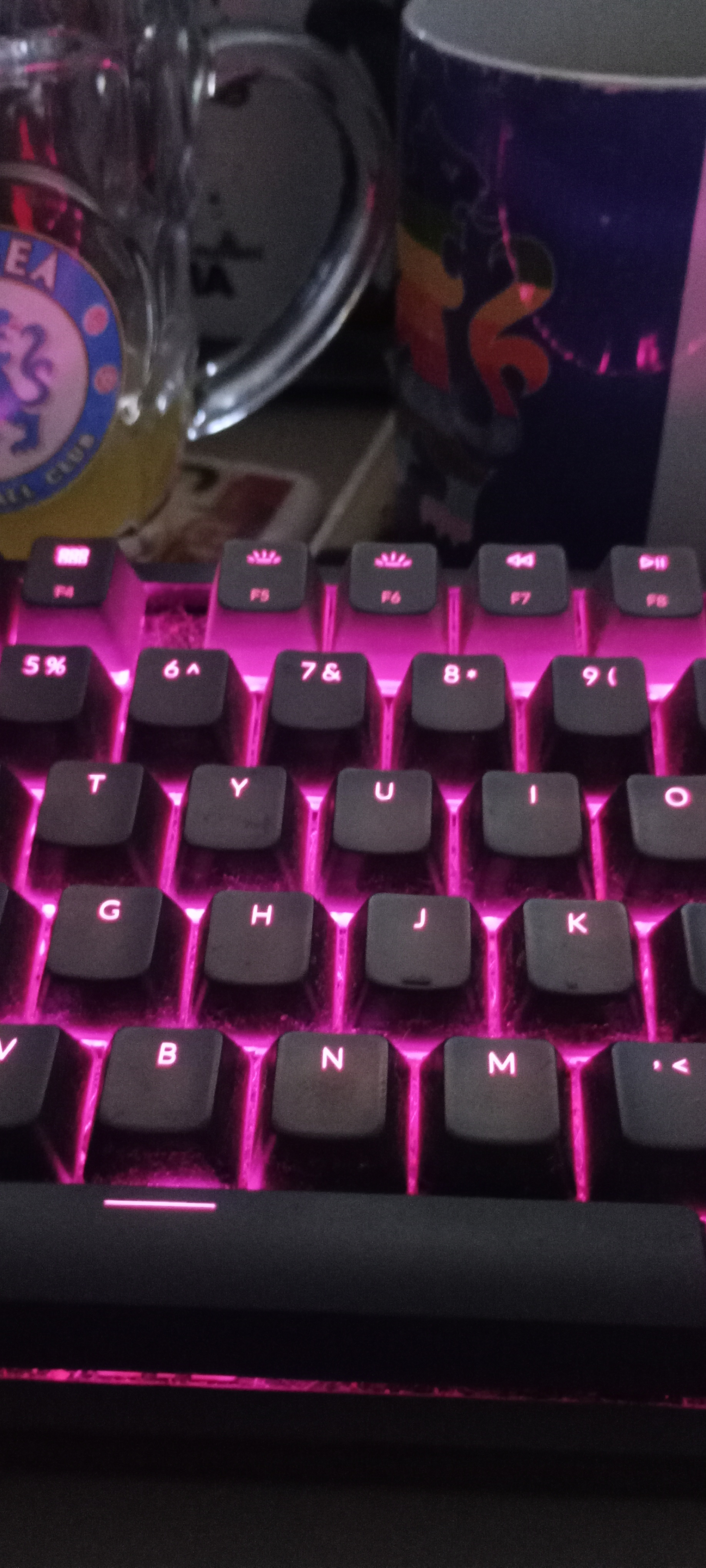 A colorful keyboard with pink lighting is placed beside two mugs, one of which has a logo on it.