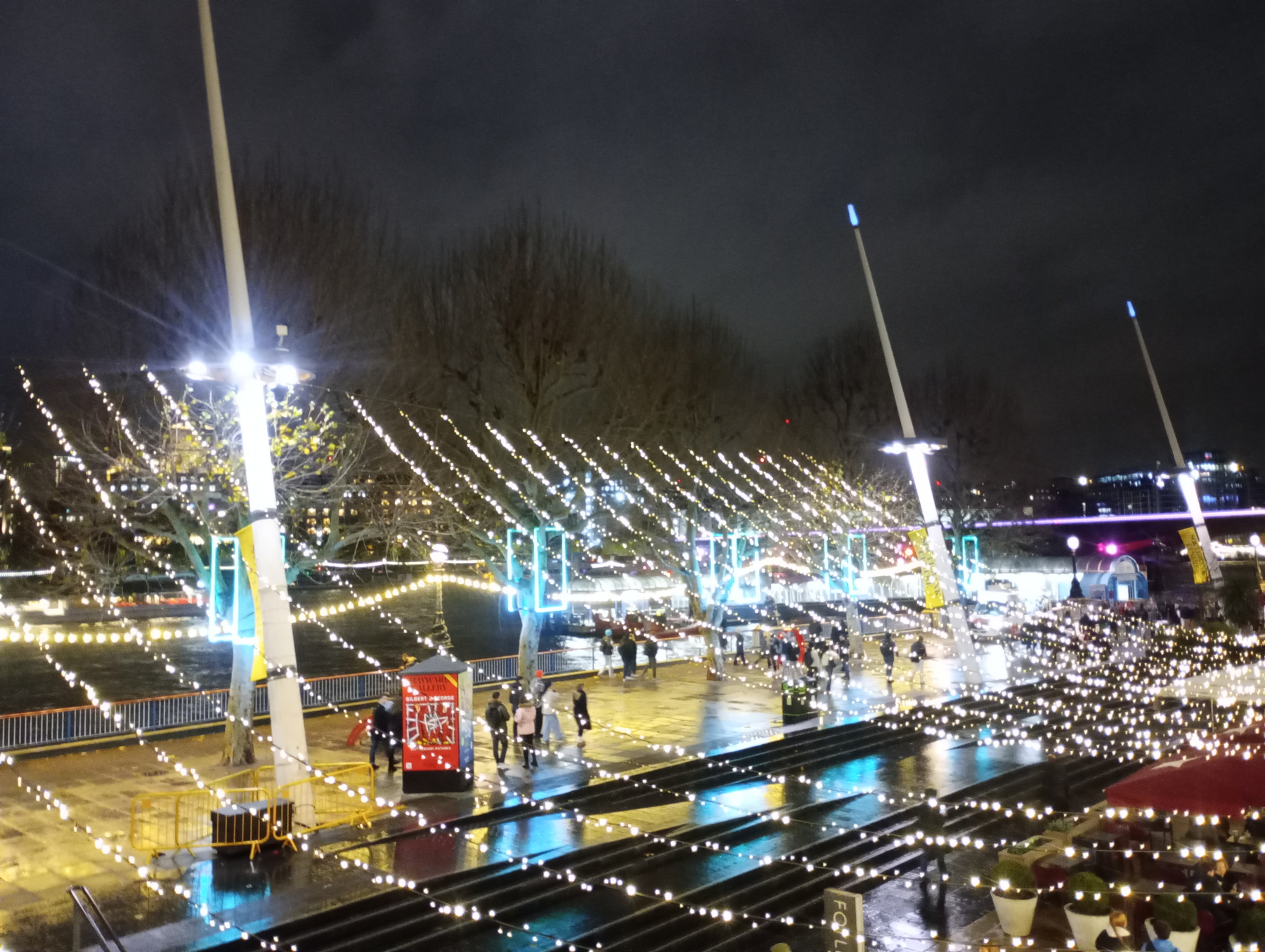 A nighttime scene with people walking under festive string lights near a waterfront area.