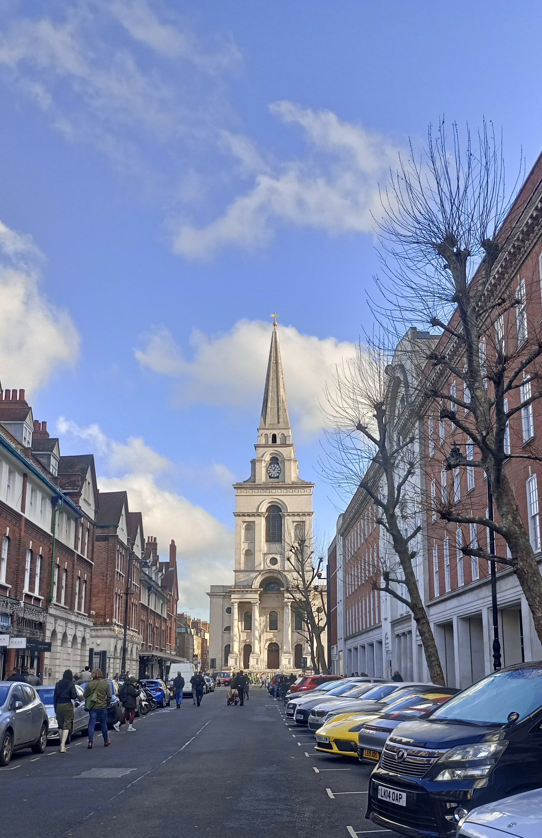 A city street lined with parked cars and red-brick buildings leads to a distant church with a tall spire under a blue sky.