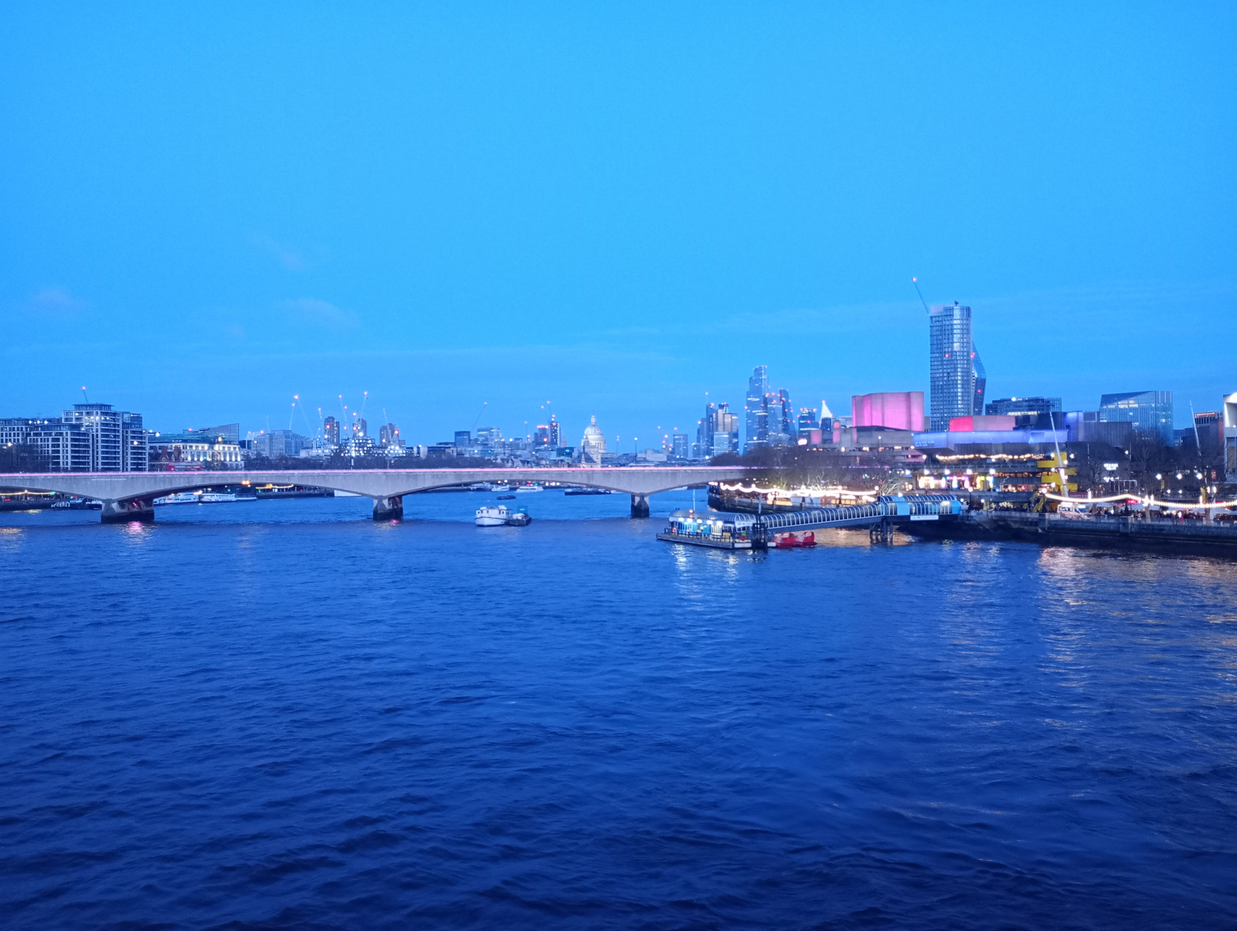 A vibrant cityscape at dusk features a river with a bridge spanning across it, surrounded by illuminated buildings and a clear blue sky.