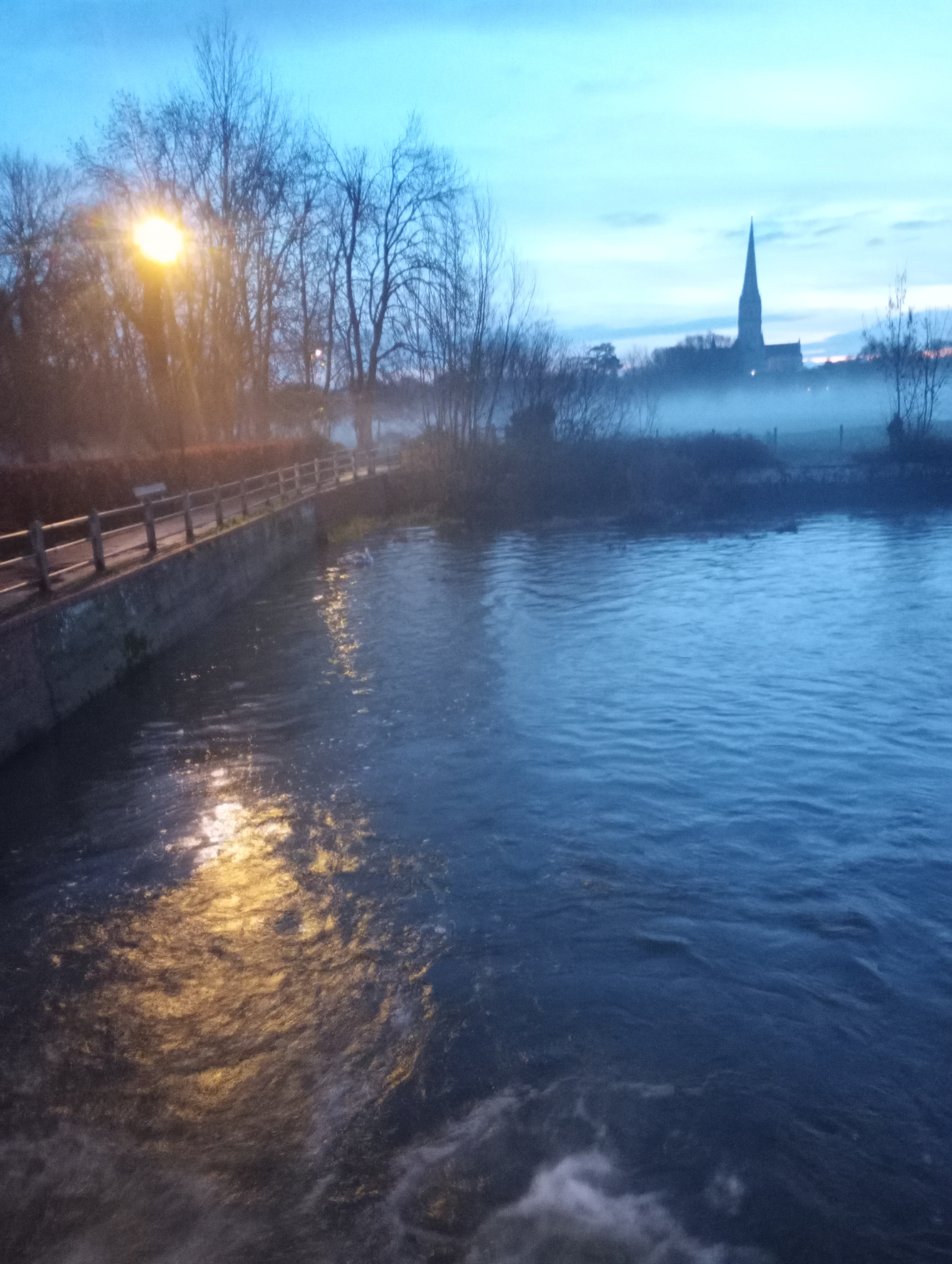 A serene evening scene features a river flowing under a bridge, with misty trees and a distant silhouette of a church spire under the twilight sky.