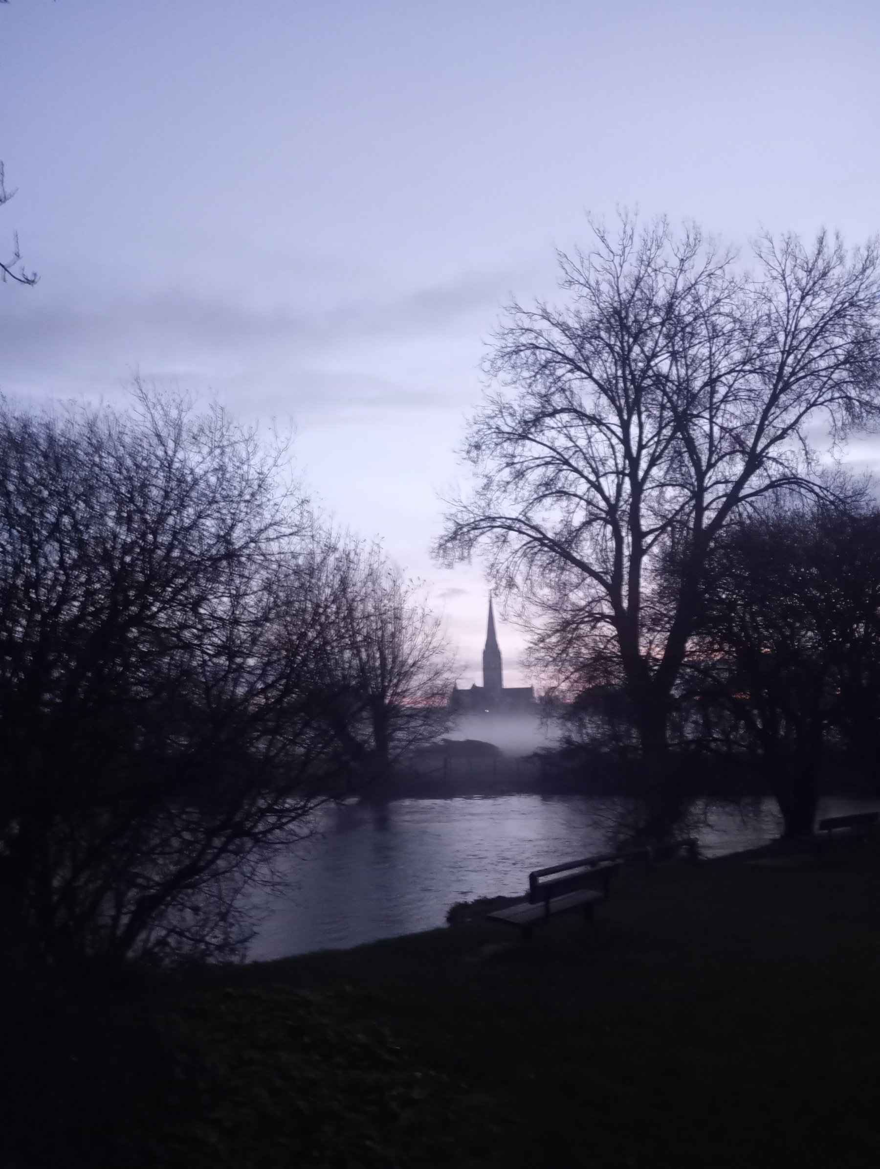A serene scene at dusk features silhouetted trees, a calm river, and a distant spire against a twilight sky.