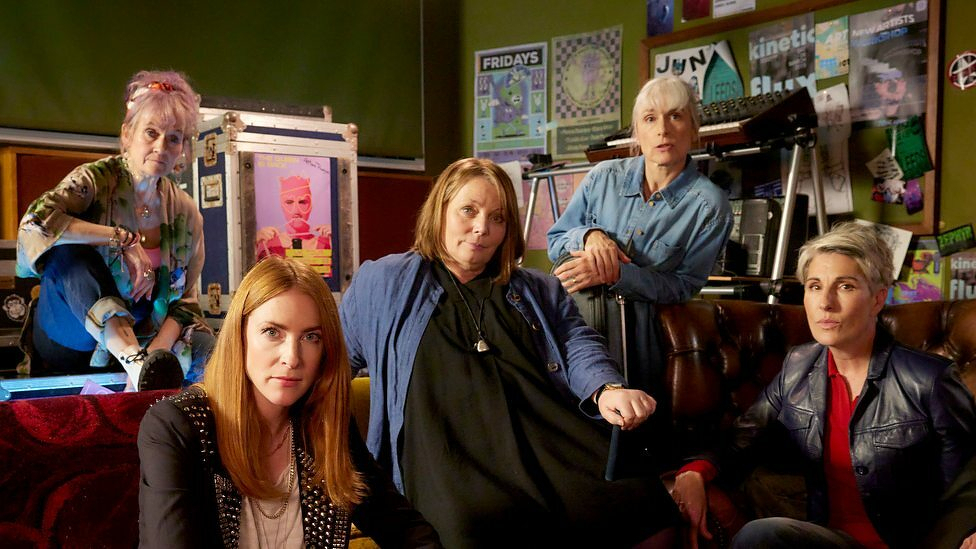 Five women are sitting and standing around a room filled with posters and props.