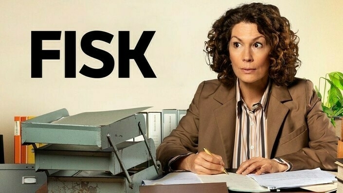 A person with curly hair, dressed in a brown suit, is sitting at a desk filled with paperwork, with the word FISK in large letters on the wall behind.
