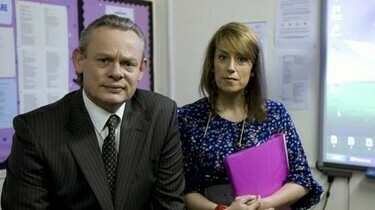 A man in a suit and a woman holding a pink folder stand in a classroom setting, with a whiteboard and bulletin board in the background.