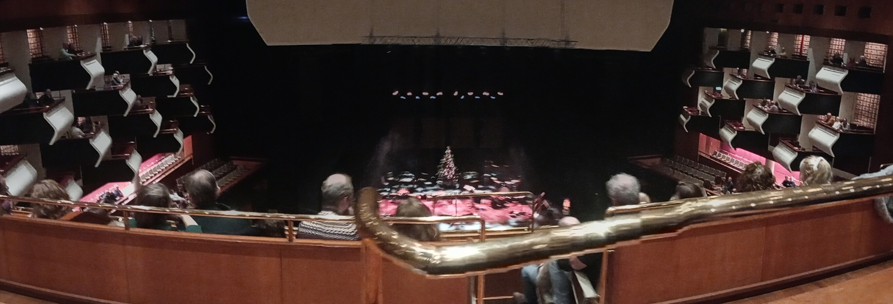 A concert hall with an orchestra on stage is viewed from an elevated seating area, featuring tiered balconies and dim lighting.