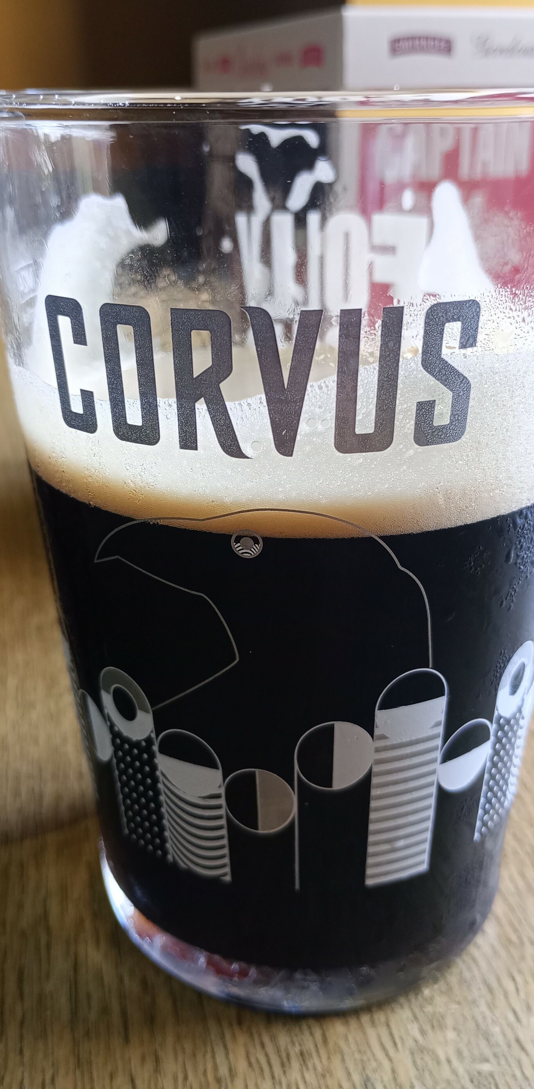 A glass filled with dark beer, featuring the word CORVUS and an artistic design on its surface, sits on a wooden table.