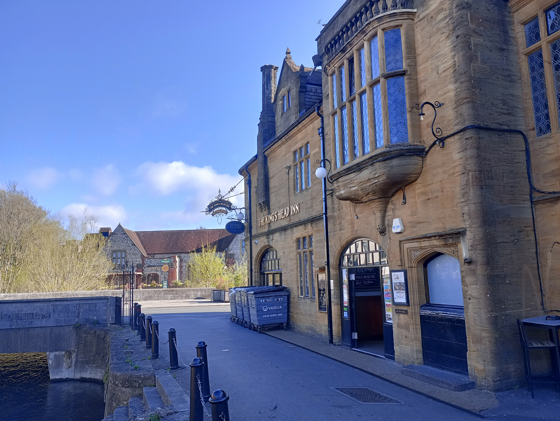 A stone building with large windows and ornate architecture is positioned along a pathway with a metal fence, adjacent to a small waterway.