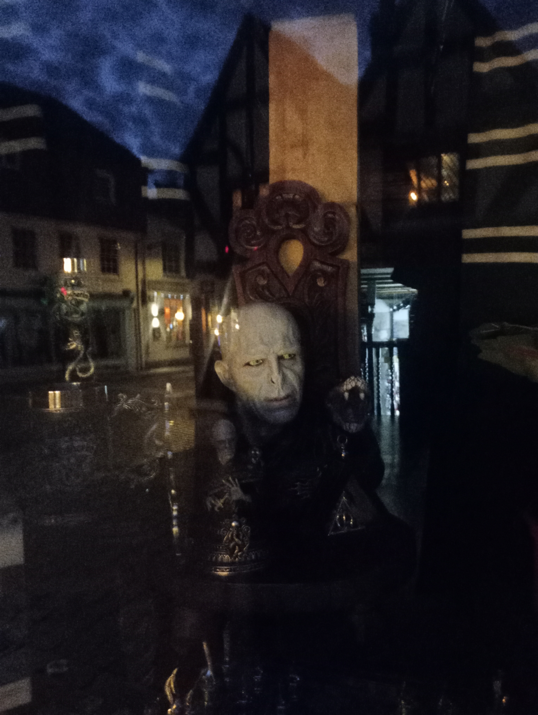 A mannequin with a pale, eerie face resembling a fantasy or horror character is displayed in a dimly lit setting with reflections of a street outside.