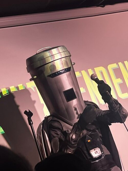 A person is dressed in a creative robot costume with a large cylindrical head and is holding a microphone while standing on a stage.