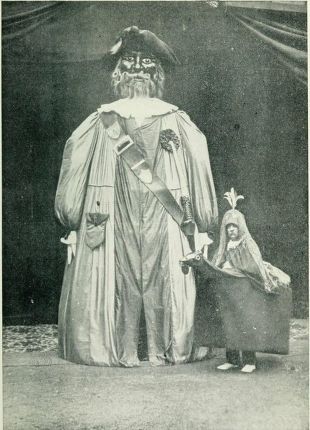 A person in a large, elaborate costume of a towering figure stands next to a child dressed in a horse costume.