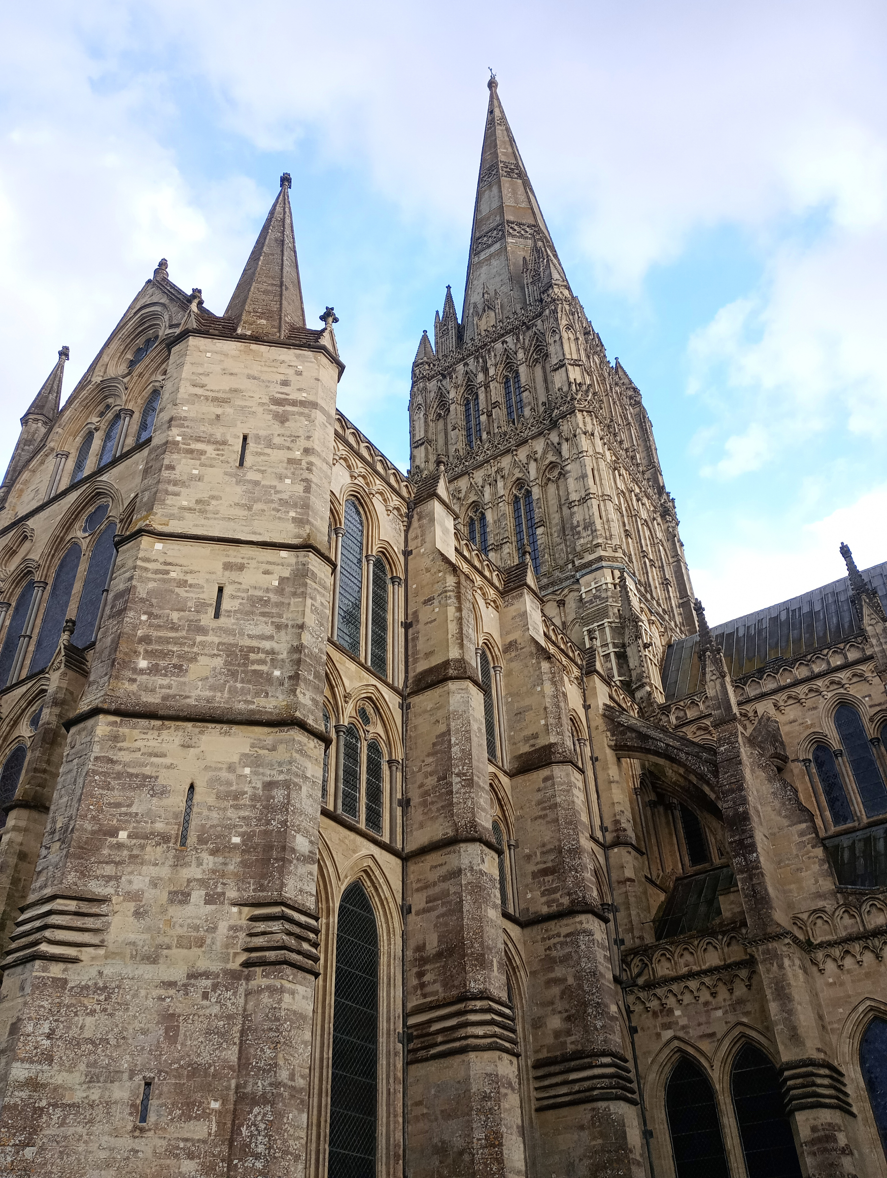A Gothic cathedral (Salisbury) with a tall spire and intricate stonework stands against a blue sky with clouds.