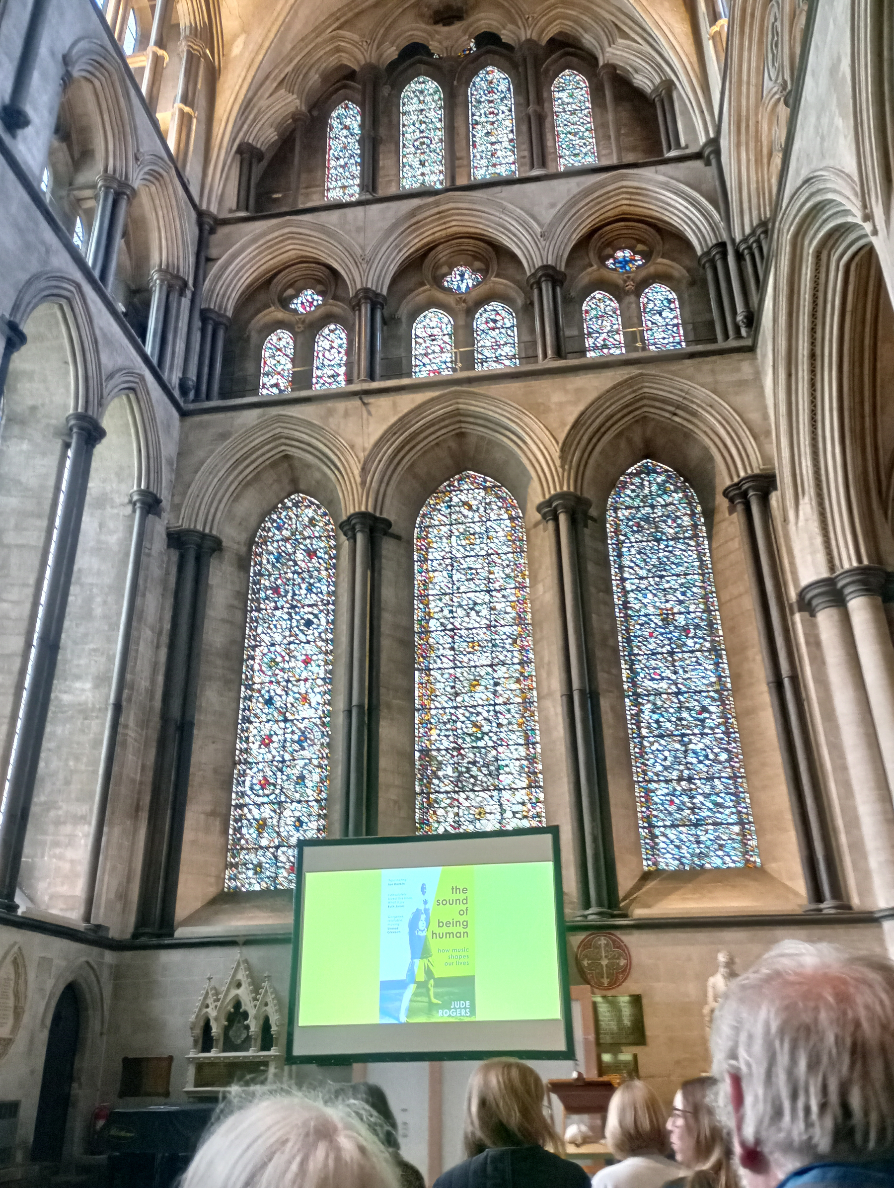 A large cathedral interior features stained glass windows and an audience watching a presentation projected on a screen.