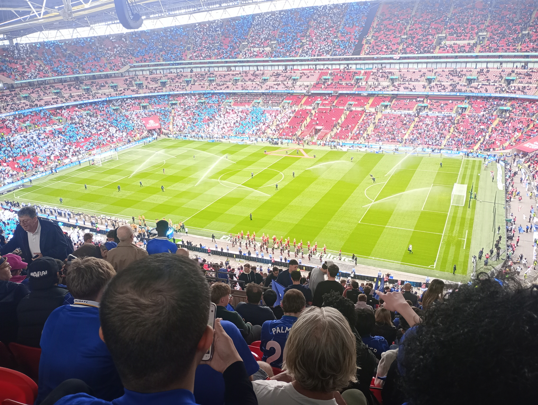 A large crowd of spectators watches a football match at a stadium with vibrant red and blue seat sections.