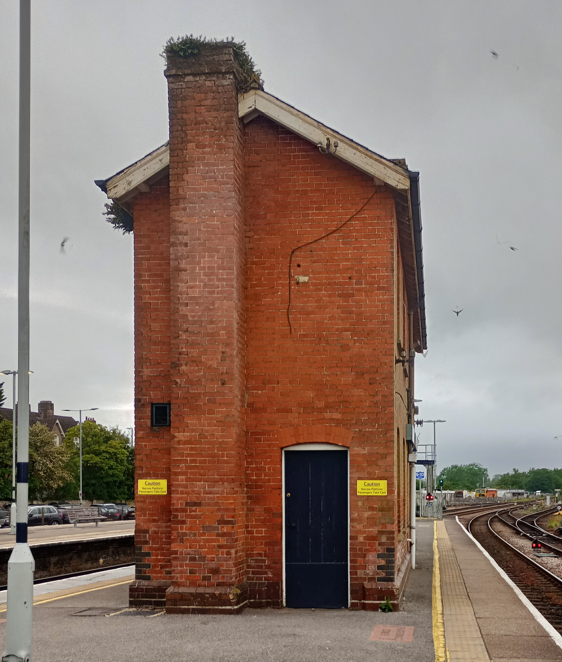 A tall, narrow brick building with a dark door stands on a railway platform under a cloudy sky.