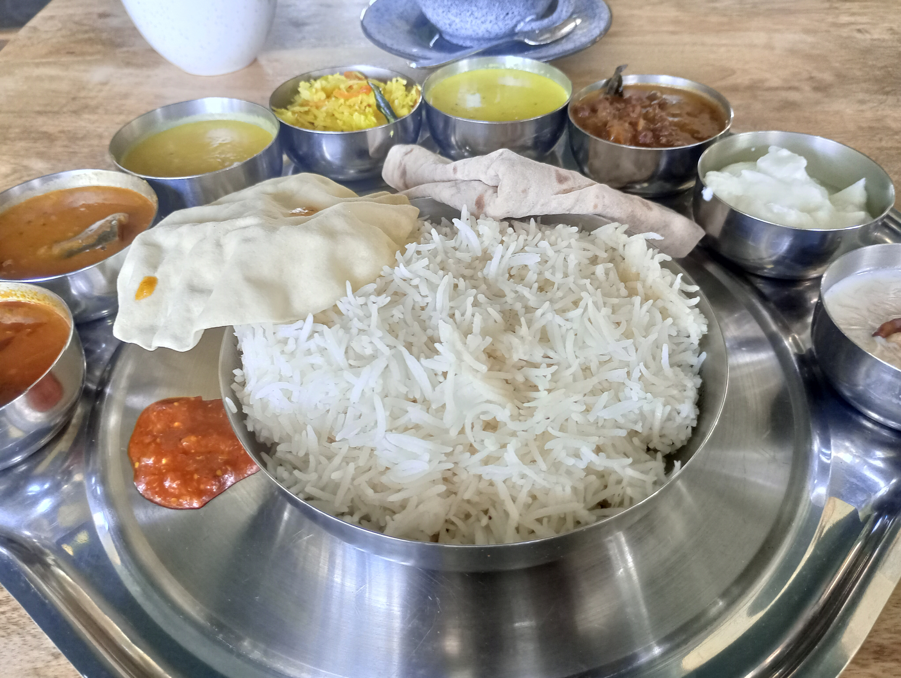 A traditional Indian thali is beautifully arranged with bowls of various curries, rice, chapati, papad, and condiments on a round metal platter.