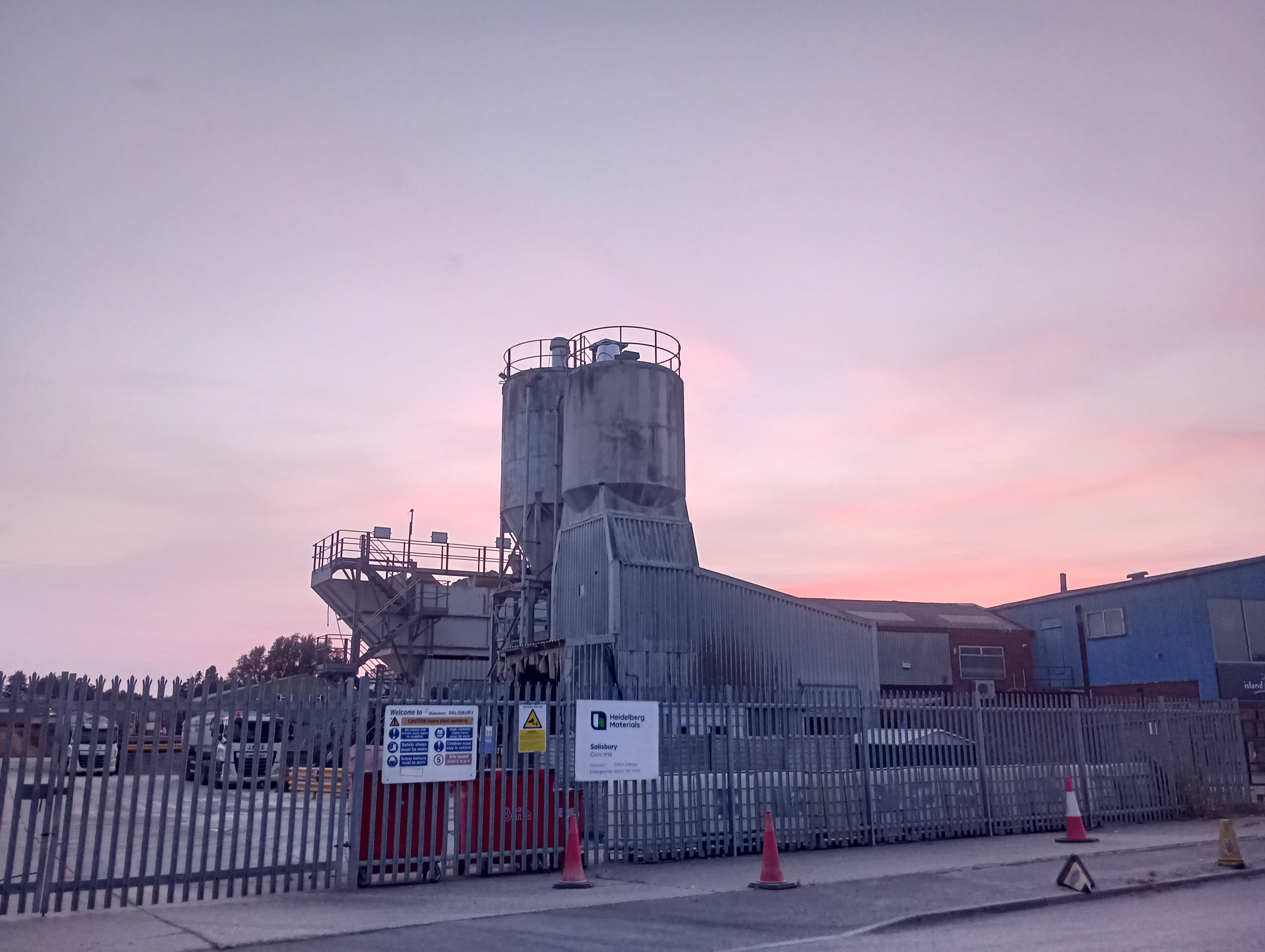 An industrial facility with large storage silos is surrounded by metal fencing and safety cones under a pink and purple sunset sky.
