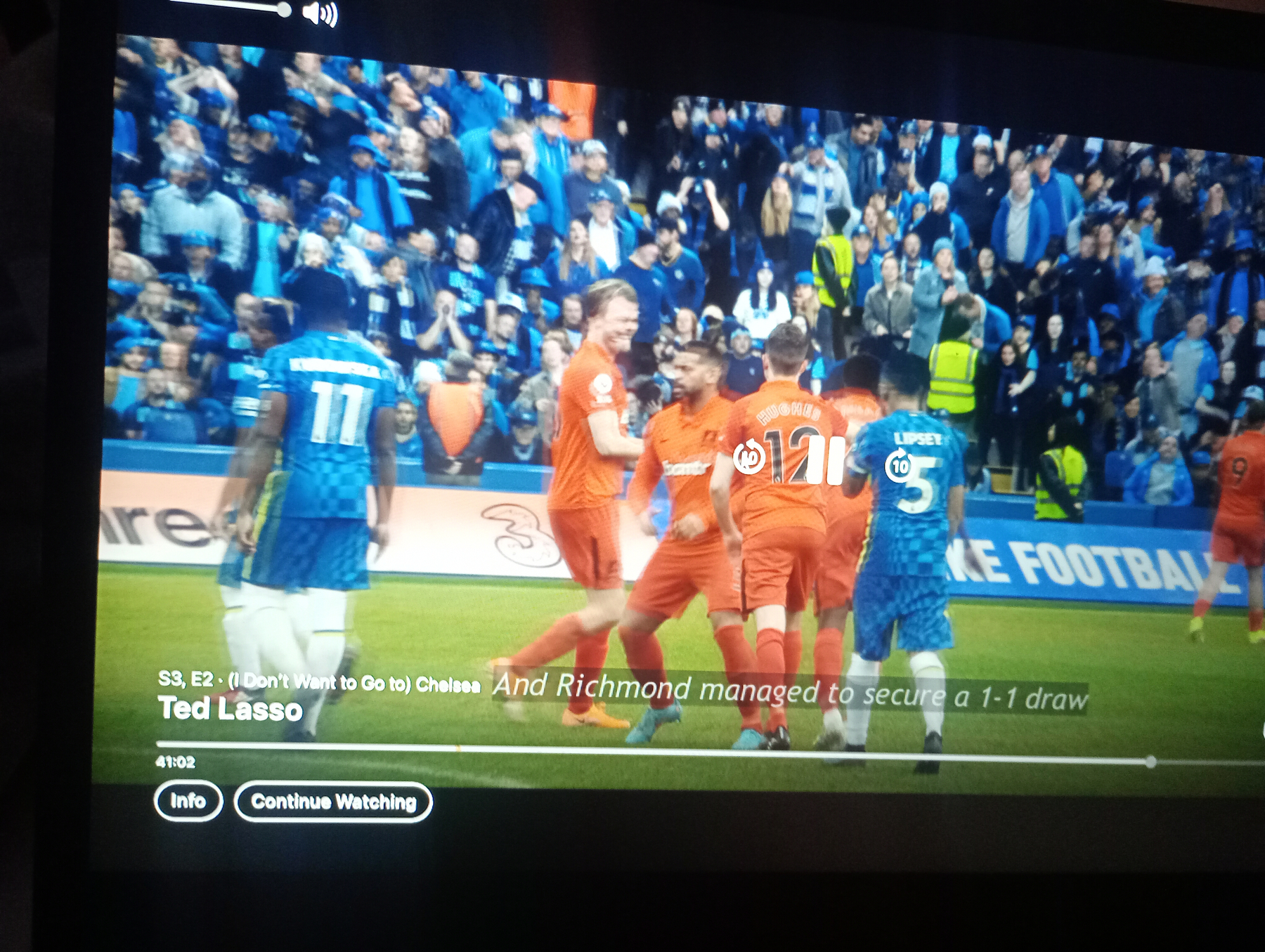 A soccer match scene from the show Ted Lasso is displayed, showing players from Chelsea and Richmond with the subtitle indicating a 1-1 draw.