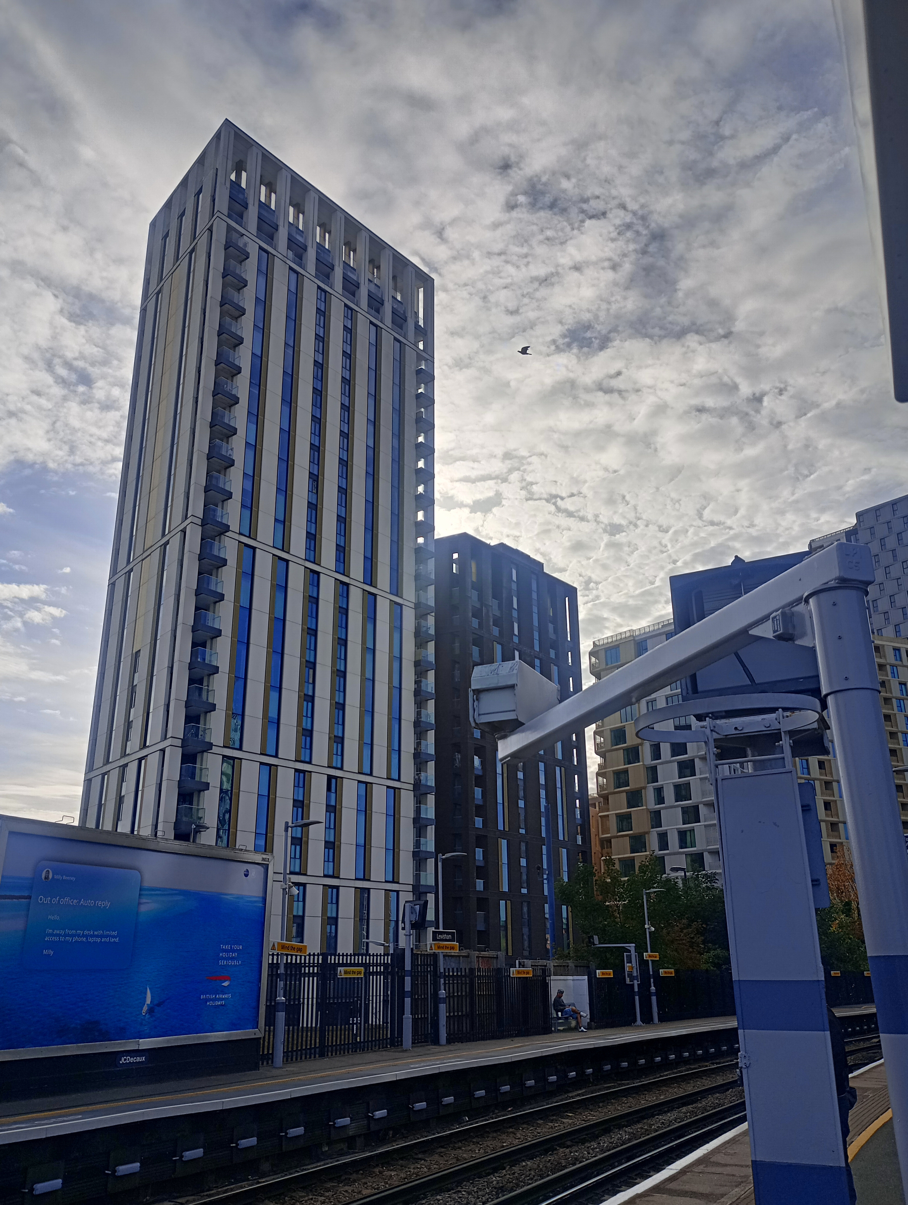 Tall modern buildings rise against a partly cloudy sky near a train station platform with tracks and a digital billboard.