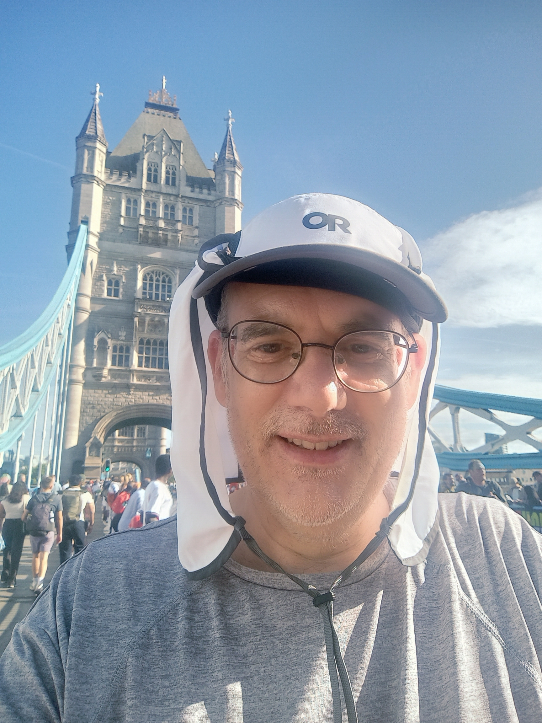 A man wearing glasses and a white cap with neck protection is smiling on a crowded pedestrian walkway of Tower Bridge in London on a sunny day.