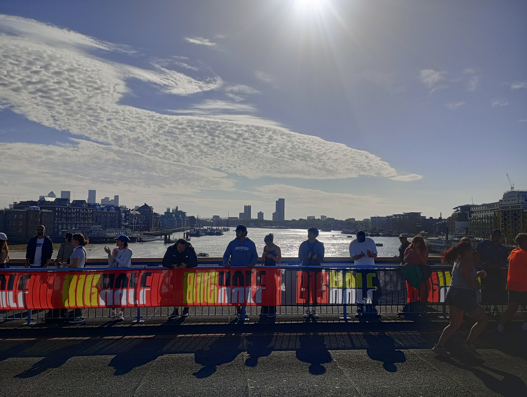 People are standing on a bridge near a colorful BRIT HALF banner with a river, city skyline, and partly cloudy sky in the background.