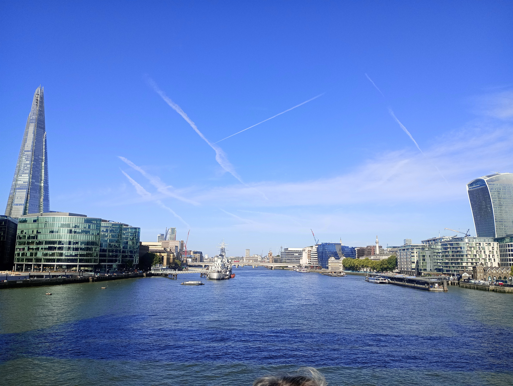 A clear blue sky stretches over the River Thames, flanked by modern buildings including The Shard on the left and the Walkie Talkie building on the right in London.